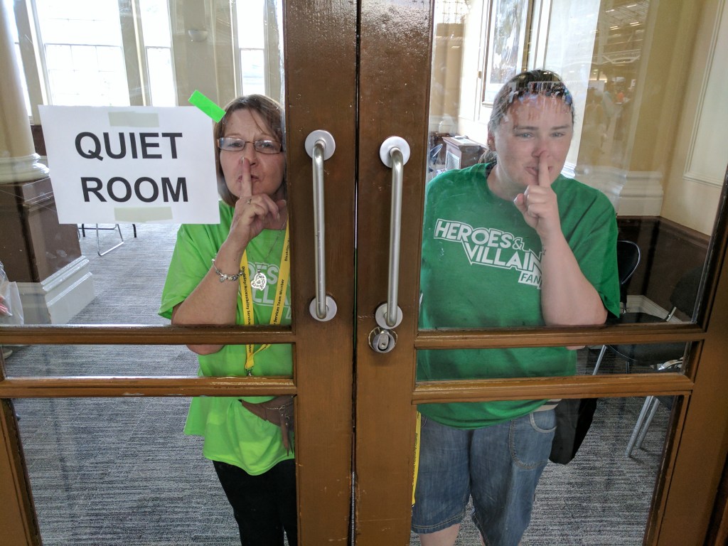 Two women in green t-shirts stand behind a pair of glass doors. A sign on the left door reads "Quiet Room" in black font.