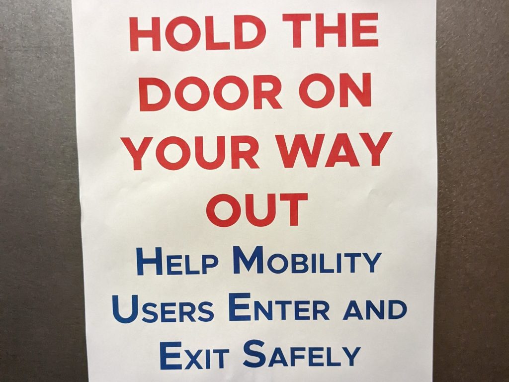 A sign reads, "Please hold the door on your way out. Help mobility users enter and exit safely." It is taped inside a gray elevator.