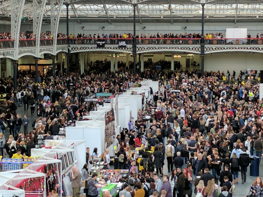 A packed event hall. A lower level is full of attendees and separated by a line of pipe-and-drape vendor booths. An upper balcony is also full of people.
