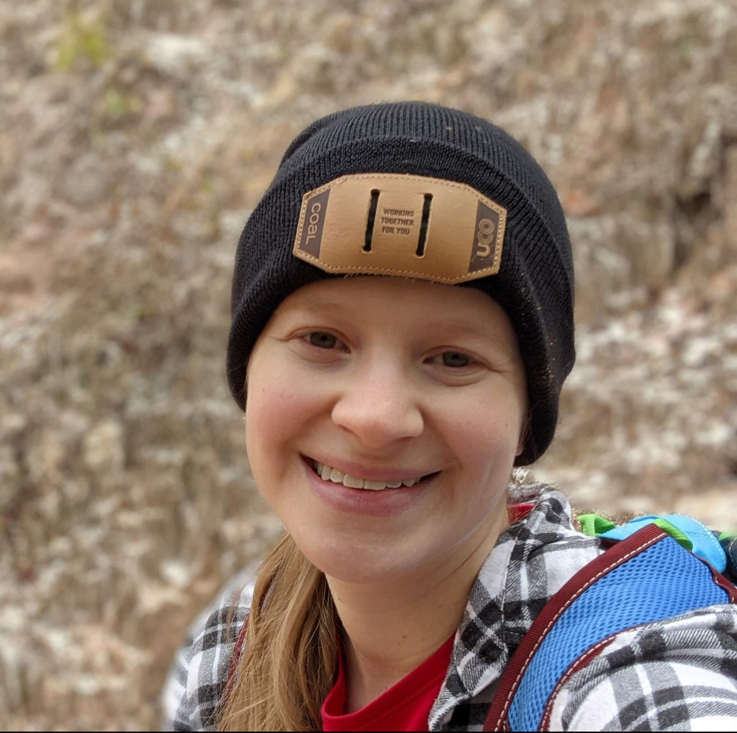 Image description: a white woman with blond hair smiles at the camera. She is wearing a black beanie and a black and white plaid shirt.